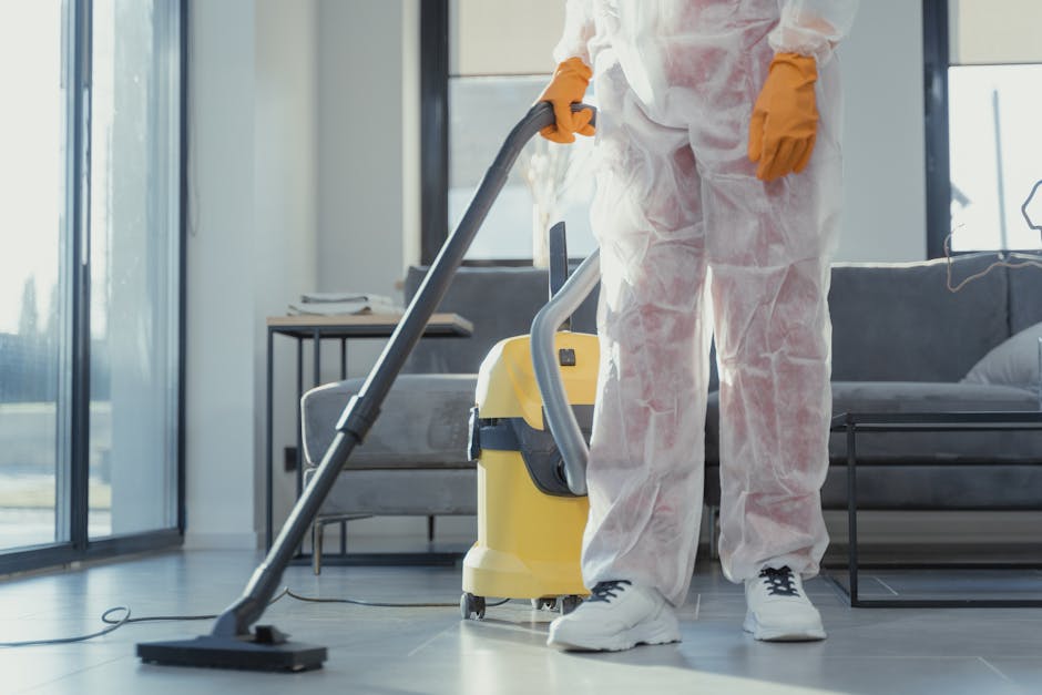 A professional cleaner wearing white protective coveralls and orange gloves operates a yellow and gray vacuum cleaner on a light gray tiled floor in a modern living room. The room features large floor