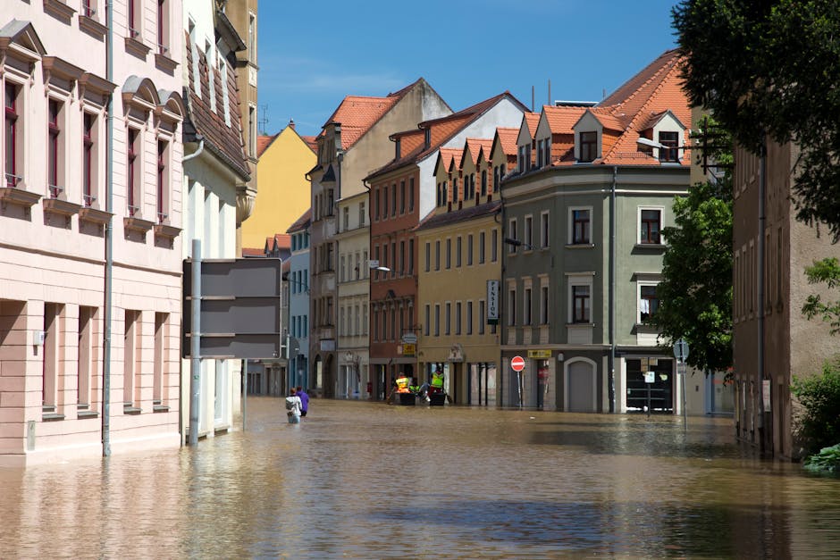 A street in Colliers Wood showing buildings with multi-colored facades, including pink, yellow, beige, and green, with red-tiled roofs. The area is heavily flooded with water covering the street surfa