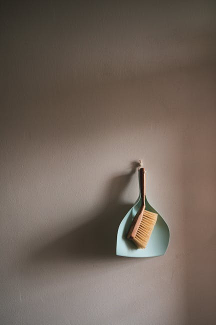 A shadowed view of a smooth, taupe-colored wall with a small hook holding a light blue, rounded-edged dish that contains a wooden-handled scrub brush and a white cleaning cloth. The dish and tools are